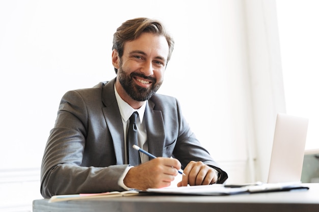 Image of laughing confident businessman wearing formal suit looking while working at laptop in office