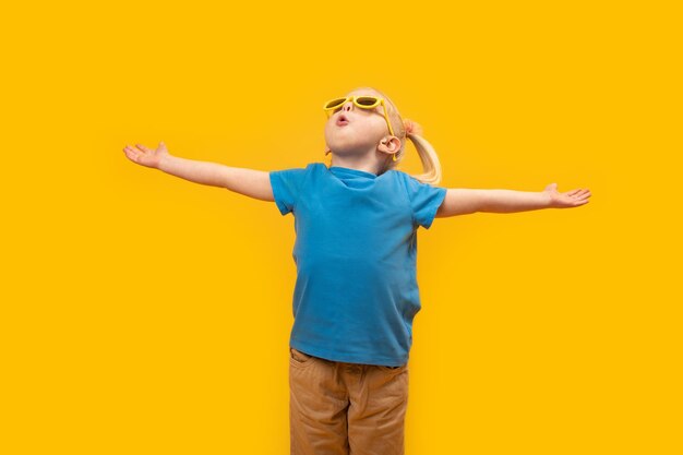 Image of little blonde girl standing isolated over yellow background wearing sunglasses and raised her hands up