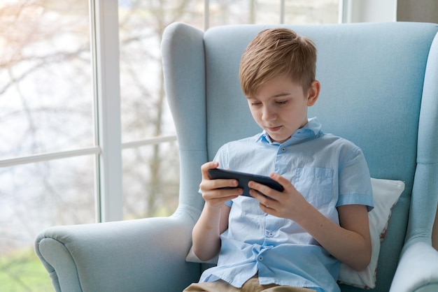 Image of little cheerful boy holding smartphone and playing game while sitting in a chair by the window at home Look at the phone Boy with the phone Cute little boy playing with smartphone