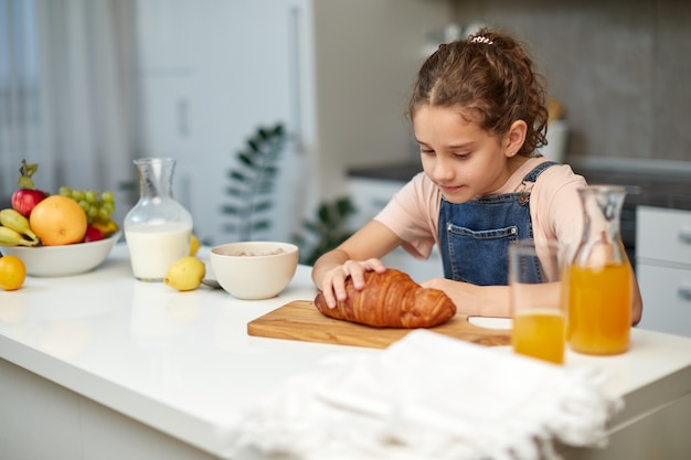 小さな巻き毛の女の子の画像は、キッチンのテーブルでクロワッサンを取ります。水平方向のビュー。