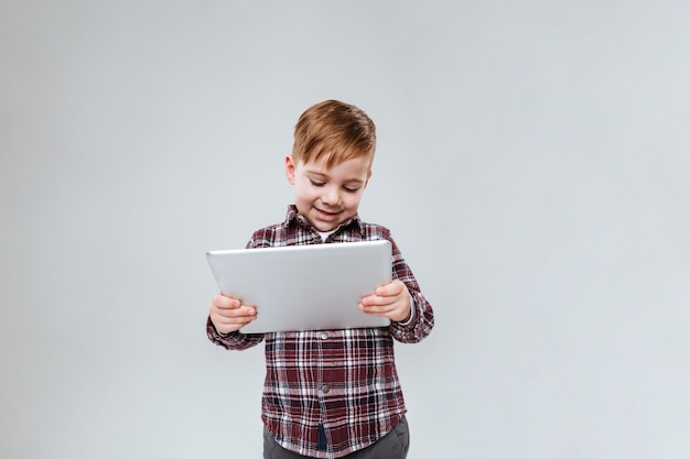 Image of little pretty child standing over grey wall holding tablet computer. Look at tablet.