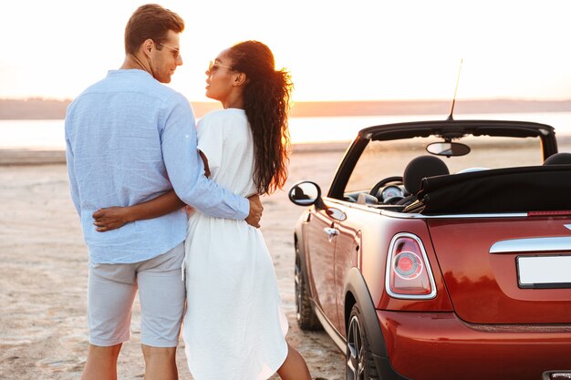 Image of lovely multiethnic couple man and woman hugging together while standing by car at seaside