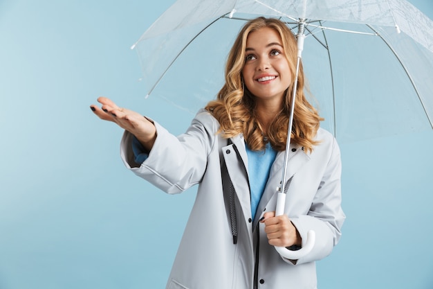 Image of lovely woman 20s wearing raincoat standing under transparent umbrella