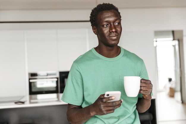 Image of masculine african american man in casual clothes holding coffee cup and smartphone at home