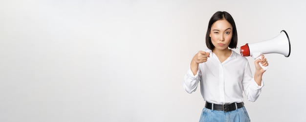 Image of modern asian woman with megaphone pointing at you camera making announcement white background