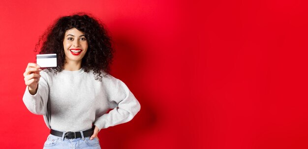 Image of modern woman with curly hair extending hand and showing plastic credit card recommending