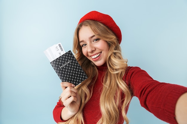 Photo image of modest blond woman 20s wearing red beret holding passport and travel ticket while taking selfie photo, isolated