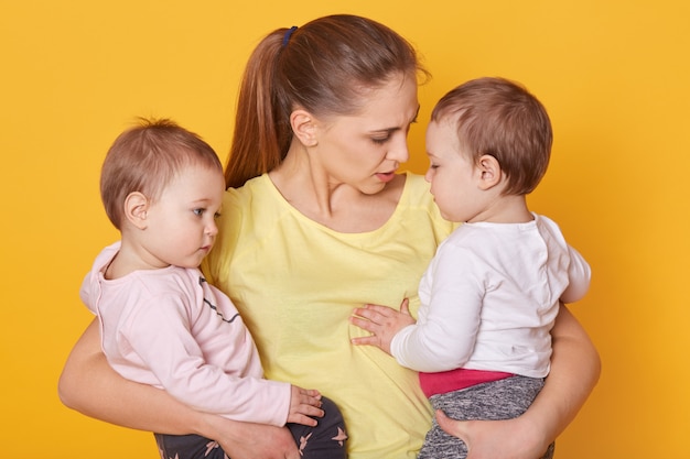 スタジオでポーズをとる彼女の甘い子供を持つ母のイメージ。カジュアルな服装のミイラと女の子の双子の母親は、母の日について彼女の娘に話します。小さな子供たちはママの話を聞いて、彼女を抱き締めます。