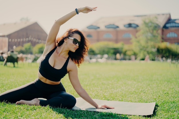 Image of motivated fit woman raises arm and does stretching\
exercises outdoors poses on fitness mat wears sunglasses and\
activewear trains actively fitness trainer goes in for sport\
outdoors