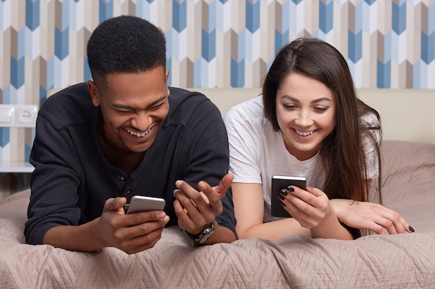 Image of multiracial couple use modern smart phones and wireless internet connection, satisfied with networking via cellular, lie in bed