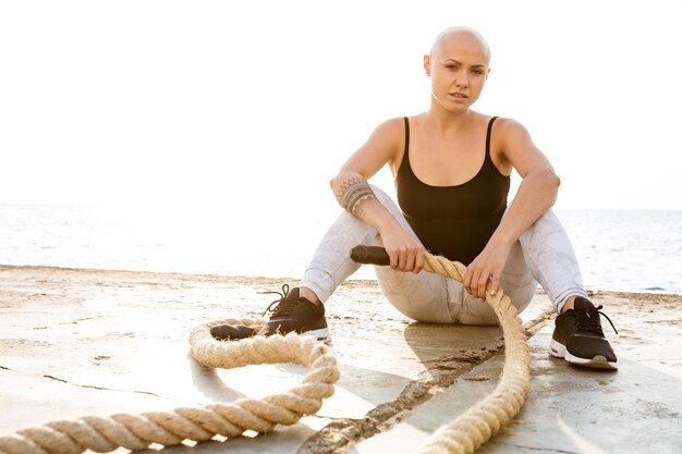 Image of nice bald woman in sportswear sitting with battle robes while working out near seaside in sunny morning