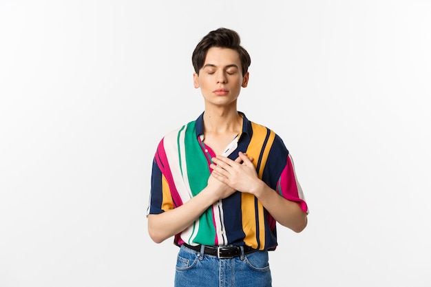 Image of nostalgic young gay man standing with closed eyes and hands on heart, remember something, standing over white