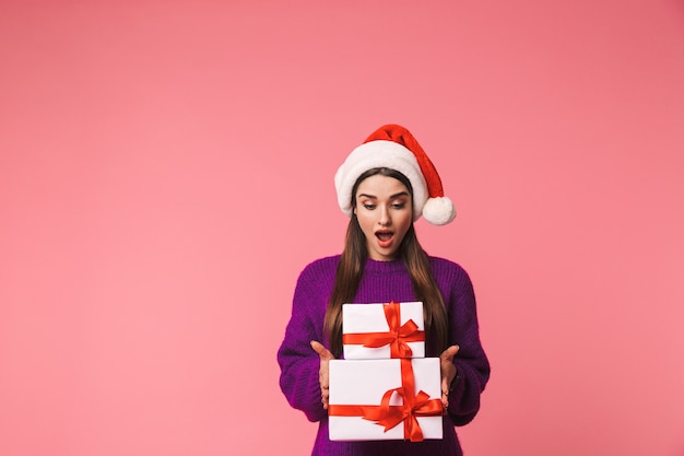크리스마스 모자를 쓰고 선물 상자를 들고 핑크 이상 격리 포즈 충격 된 젊은 감정적 인 여자의 이미지.