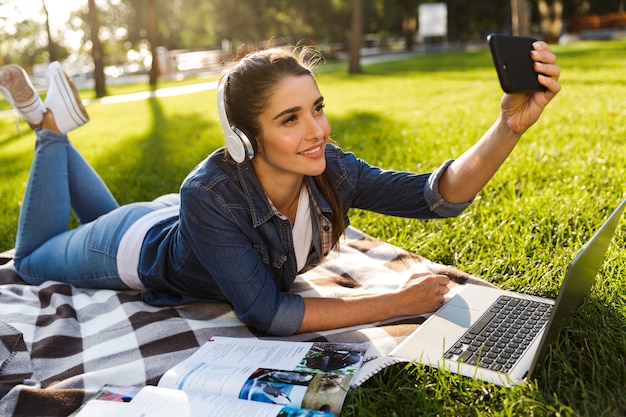 Фото Изображение удивительной красивой молодой женщины-студента в парке с помощью портативного компьютера, слушающего музыку в наушниках, делает селфи по телефону.