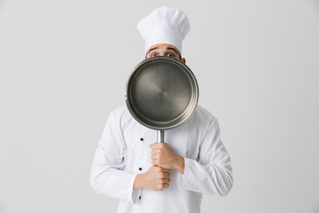 Фото Изображение возбужденного эмоционального шеф-повара молодого человека в помещении изолированного над предпосылкой белой стены держа сковороду.