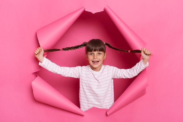 Фото Изображение счастливой довольной забавной маленькой девочки в полосатой рубашке, позирующей в порванных розовой бумаге поднятых руках с косичками в руках, смотрящей в камеру с позитивным выражением лица