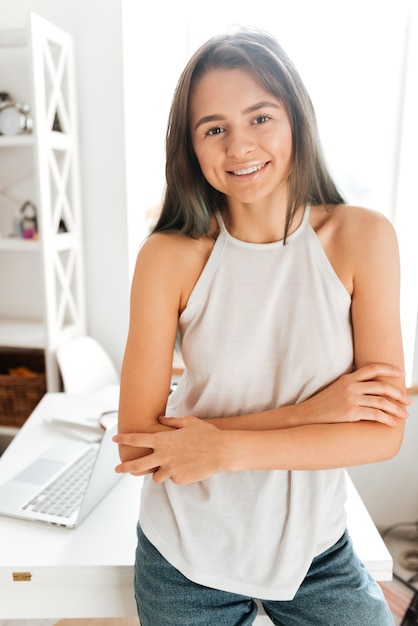 写真 テーブルの近くで腕を組んで家に立っているかなり若い女性の画像。カメラを見てください。