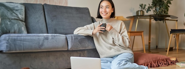 写真 カップを持って熱いお茶を飲み、床のノートパソコンの近くに座って休んでいる笑顔のアジア人女性の画像