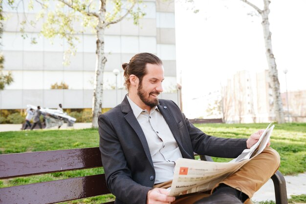 Изображение стильного красавца 30-х в деловом костюме, сидящего на скамейке в зеленом парке и читающего газету в солнечный день