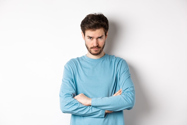 Image of offended and sad young man with beard, look from under forehead and sulking irritated, cross arms on chest defensive, mad at someone, standing on white background