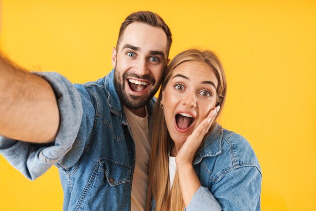 Image of optimistic couple man and woman in denim clothes smiling  while taking selfie photo isolated over yellow wall