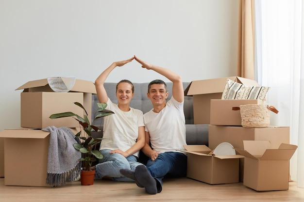Изображение оптимистичной пары в белых футболках в стиле кэжуал, позирующей с картонными коробками во время переезда в новую квартиру, создающей крышу над головой в безопасности