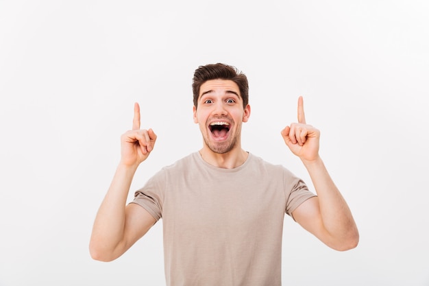 Image of optimistic man in casual t-shirt smiling and pointing fingers upwards on copyspace text or product, isolated over white wall