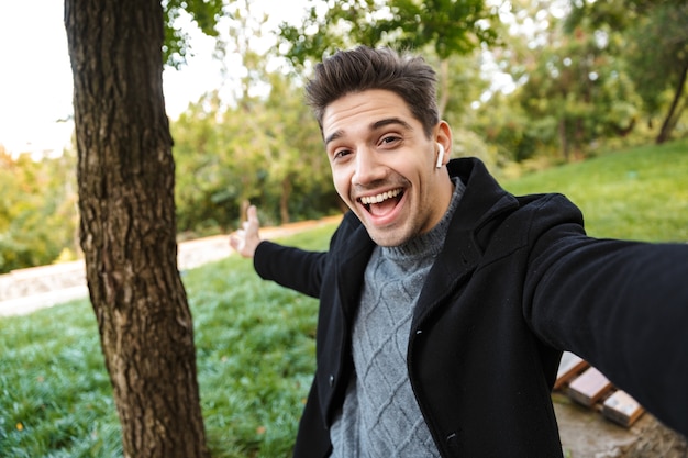 Image of optimistic pleased young man in casual clothing walking outdoors in green park take a selfie  listening music with earphones showing copyspace.