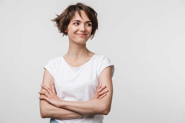 Image of optimistic woman in basic t-shirt smiling and looking aside while standing with arms crossed isolated over white wall