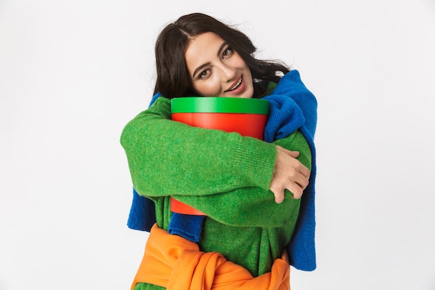 Image of optimistic woman with dark hair in colorful clothes holding big gift box while standing, isolated on white