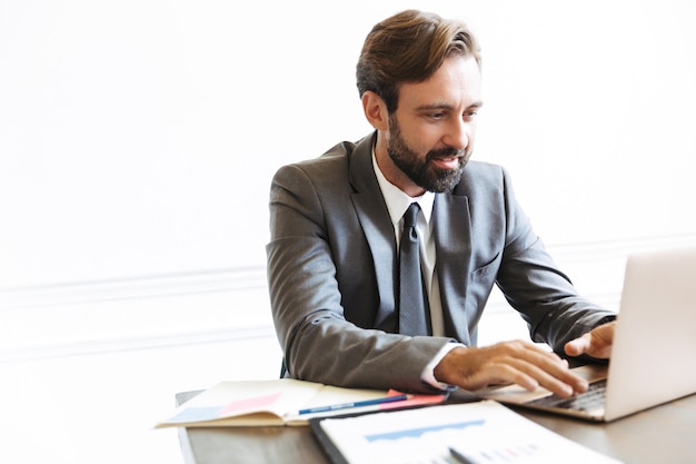 Image of optimistic young pleased business man sitting in office using laptop computer working indoors.