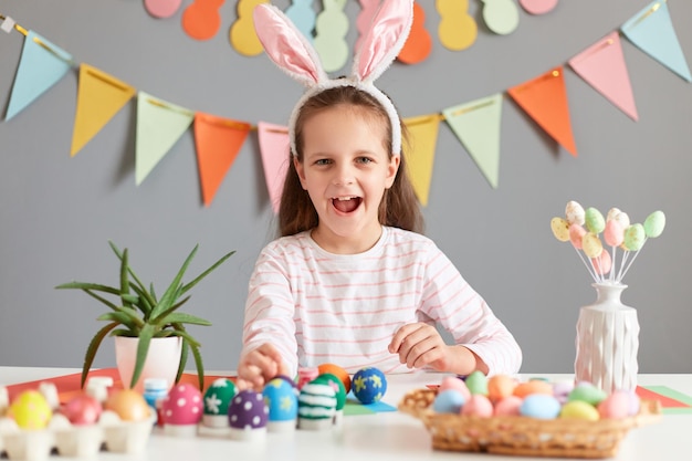 Image of overjoyed cheerful little girl sitting at table among multicolored painted dyed easter eggs wearing bunny ears preparing for celebration yelling with positive expression