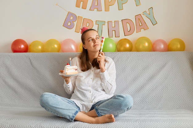 Image of pensive adorable young adult woman wearing white shirt celebrating birthday sitting alone on cough with cake holding party horn looking away dreaming