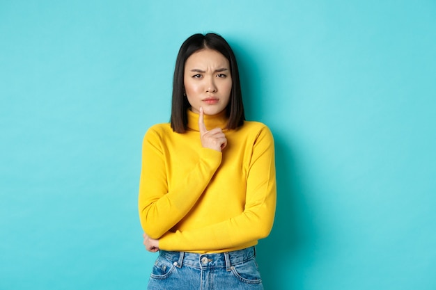 Image of pensive asian woman touching lip and frowning, thinking about something, trying to understand, standing over blue background.