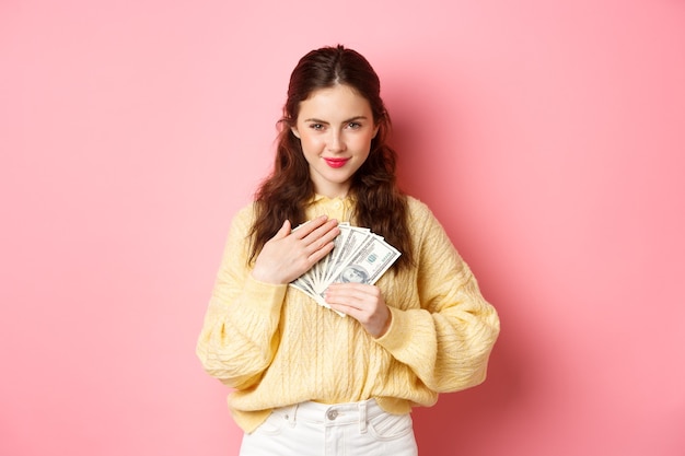 Immagine di una donna bruna contenta che tiene soldi vicino al petto, abbraccia banconote da un dollaro e guarda timidamente la telecamera con un sorriso sicuro, in piedi su sfondo rosa.