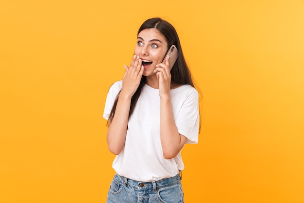 Image of pleased brunette woman wearing basic clothes rejoicing while talking on cellphone isolated over yellow wall