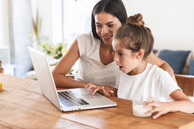 Image of pleased family woman and her little daughter smiling and using laptop computer together while sitting at table in apartment