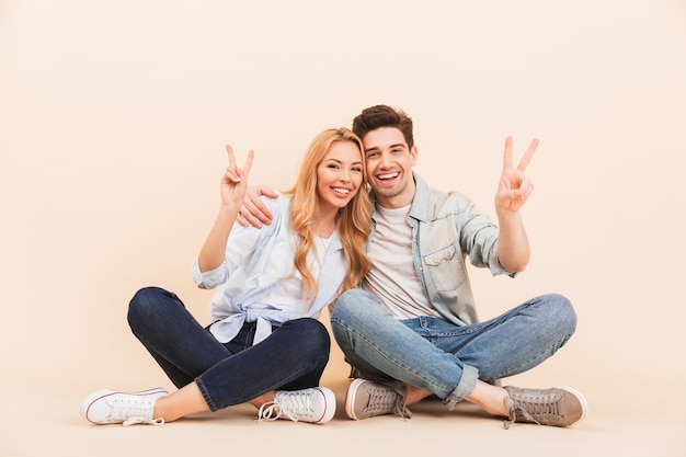 Image of pleased man and woman hugging together while sitting on the floor with legs crossed and showing peace symbols, isolated over beige wall