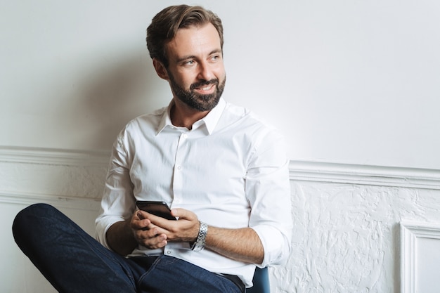 Image of pleased smiling businessman wearing white shirt typing on cellphone and looking aside while working in office