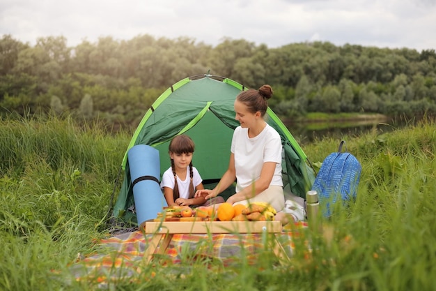 Immagine di una madre felice e felice positiva e sua figlia che fanno un picnic vicino al fiume seduti su una coperta per terra e parlano divertendosi a trascorrere del tempo insieme