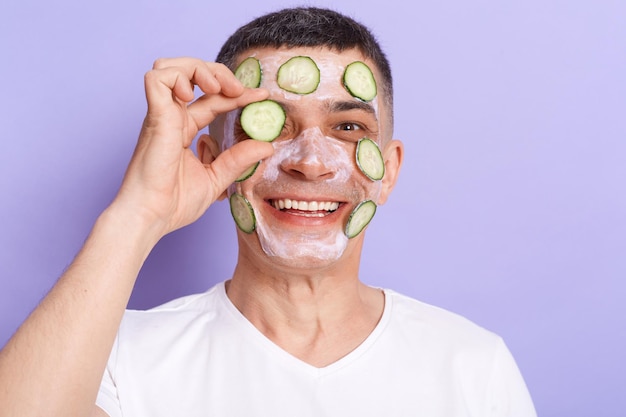 Image of positive joyful man wearing white t shirt applying mask posing isolated over purple background covering hid eye with cucumber slice looking at camera with toothy smile