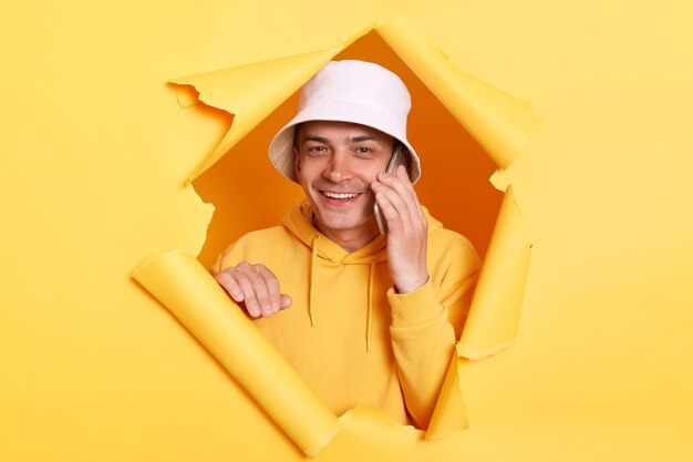 Image of positive optimistic man wearing hoodie and panama posing in hole in yellow paper wall standing with happy expression and talking via smart phone hearing good news