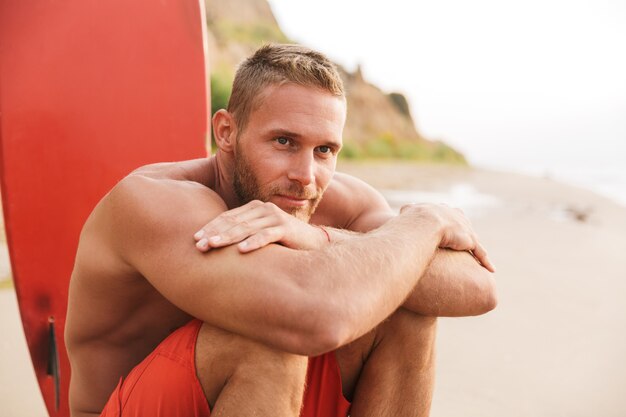 Photo image of a positive strong handsome man surfer with surfing on a beach outside.