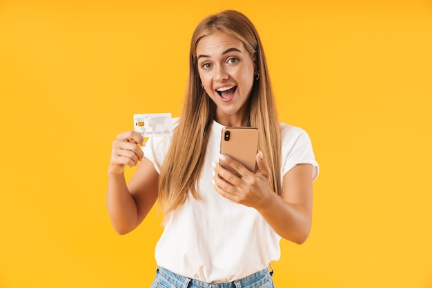 Image of positive woman in basic clothes smiling while holding smartphone and credit card isolated over yellow wall