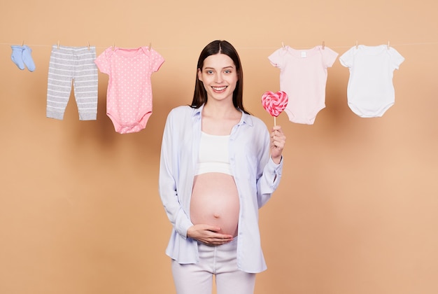 Image of pregnant joyful young Caucasian girl standing and posing with lollipop on pink background. The concept of motherhood, childbirth. Pregnancy, women and beauty concept.