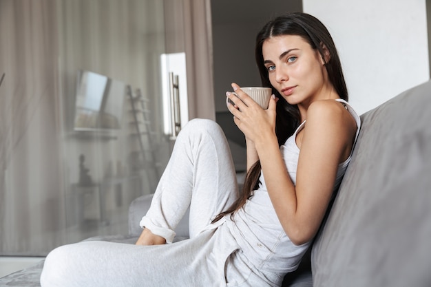 Image of pretty beautiful young woman sitting on sofa indoors at home drinking coffee or tea.