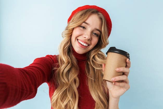 Photo image of pretty blond woman 20s wearing red beret holding paper cup with coffee while taking selfie photo, isolated