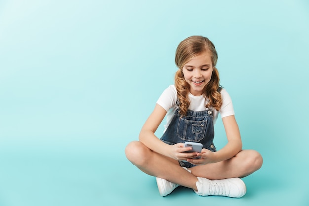 Image of pretty happy young little girl posing isolated over blue wallusing mobile phone chatting.