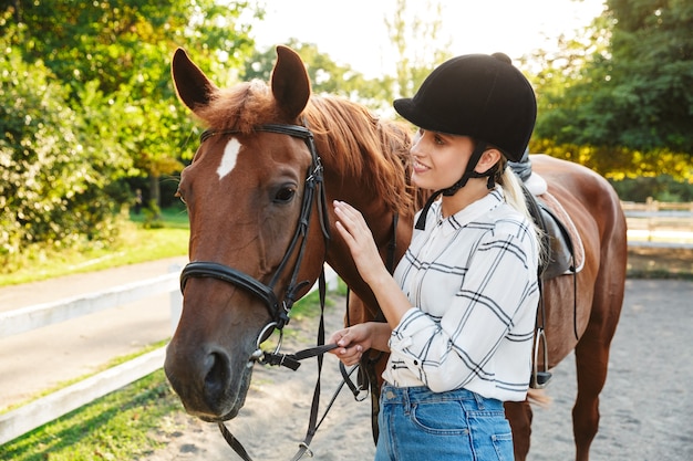 田舎の庭で馬のそばに立っている帽子をかぶっているかなり若いブロンドの女性の画像