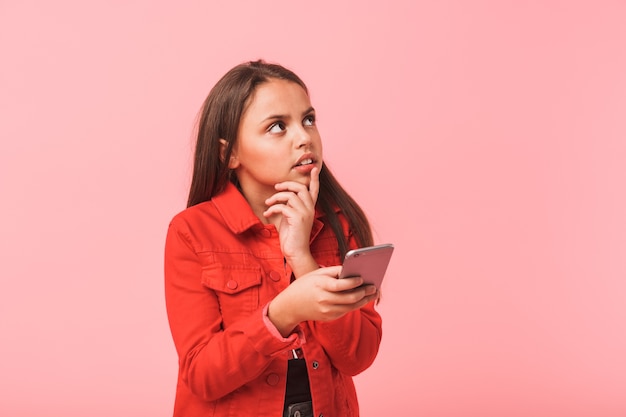 Image of puzzled teen girl in casual using cell phone while standing, isolated over red wall
